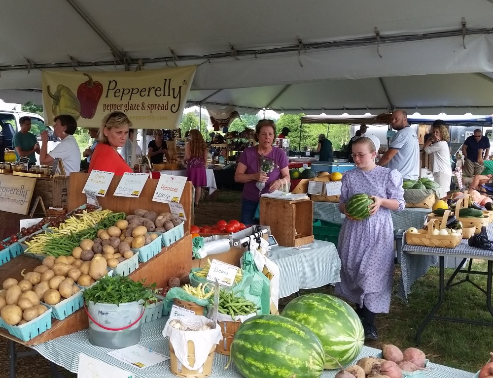 The inside of a tent at the Farmers Market in Hershey, with a table full of produce in the foreground and marketgoers browsing through produce and other goods at tables in the background.