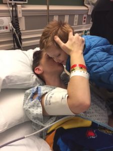 A young boy leans over a woman in a hospital bed as she holds his head with her right arm and kisses him on the cheek.