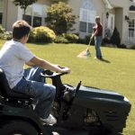 A teen sits on a riding mower in the front yard of a home. He looks uphill toward the house and a man raking grass.