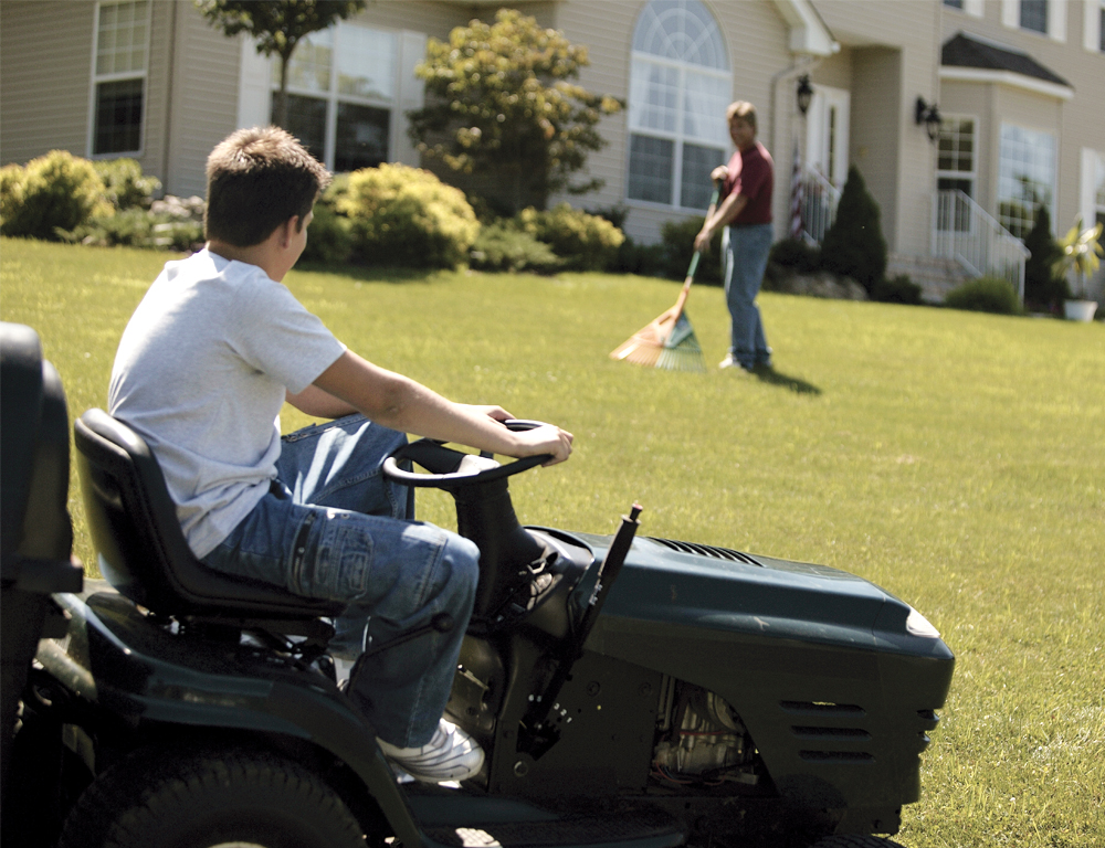 A teen sits on a riding mower in the front yard of a home. He looks uphill toward the house and a man raking grass.