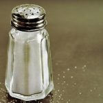 : A close-up of a full salt shaker sitting on a counter. A few grains of salt are sprinkled around it.