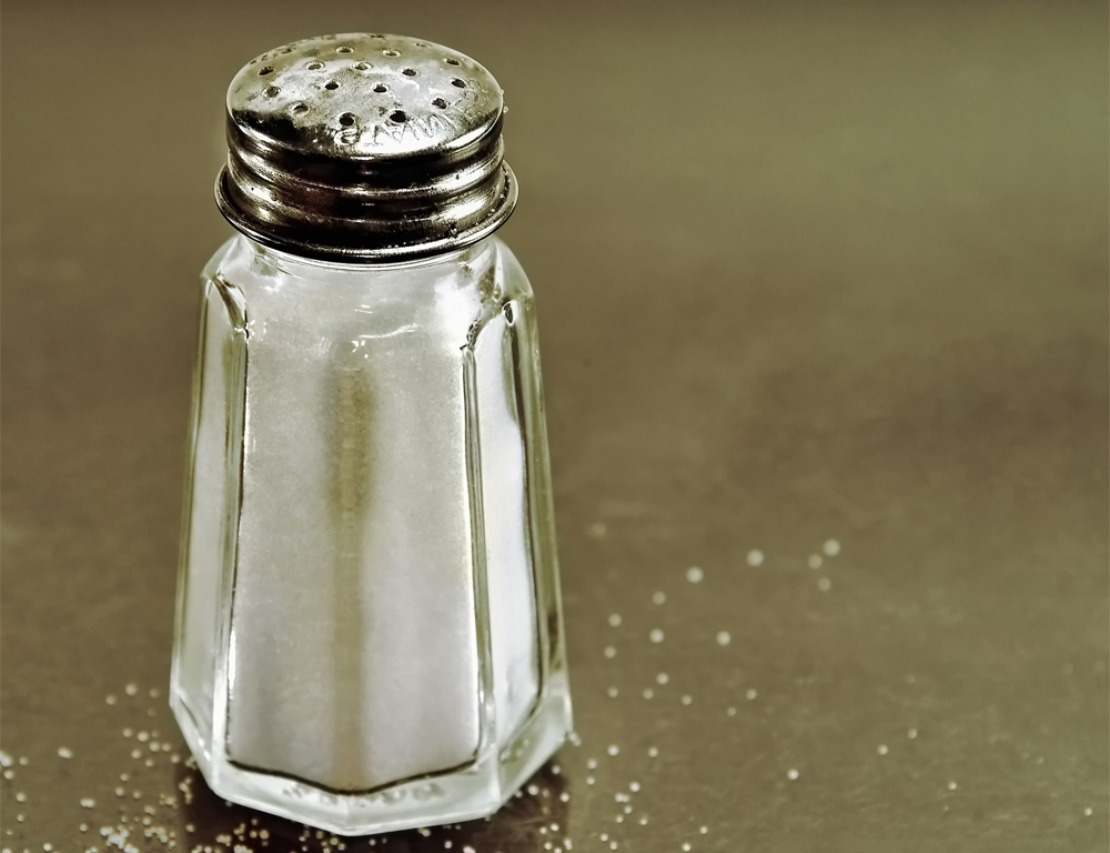 : A close-up of a full salt shaker sitting on a counter. A few grains of salt are sprinkled around it.