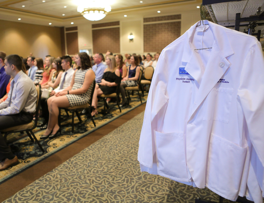 A white physician assistant coat is hung in the foreground, to the right. In the background, to the left, several people are seated in a small hotel ballroom.