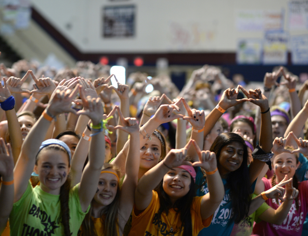 Several young people in a school gymnasium hold their arms in the air, making a diamond formation using the thumb and forefinger of each hand.