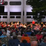 A crowd is in the foreground, seated on lawn chairs and blankets, with an orchestra in the background. The backdrop is Hershey Medical Center's signature "Crescent."
