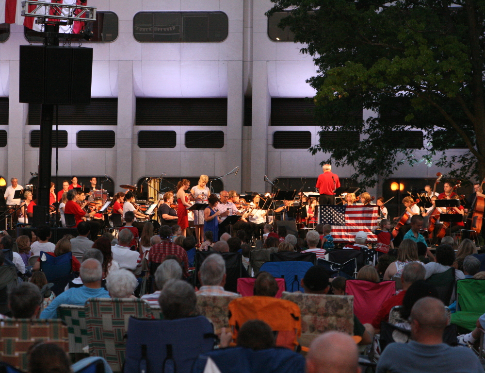 A crowd is in the foreground, seated on lawn chairs and blankets, with an orchestra in the background. The backdrop is Hershey Medical Center's signature 