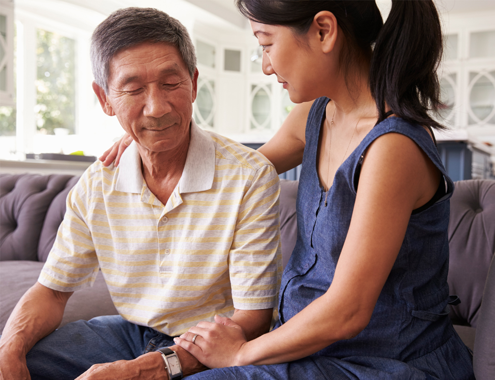 A young woman sits on a couch next to an older man. She has her right arm around him and looks at him. He has a neutral expression and looks downward.