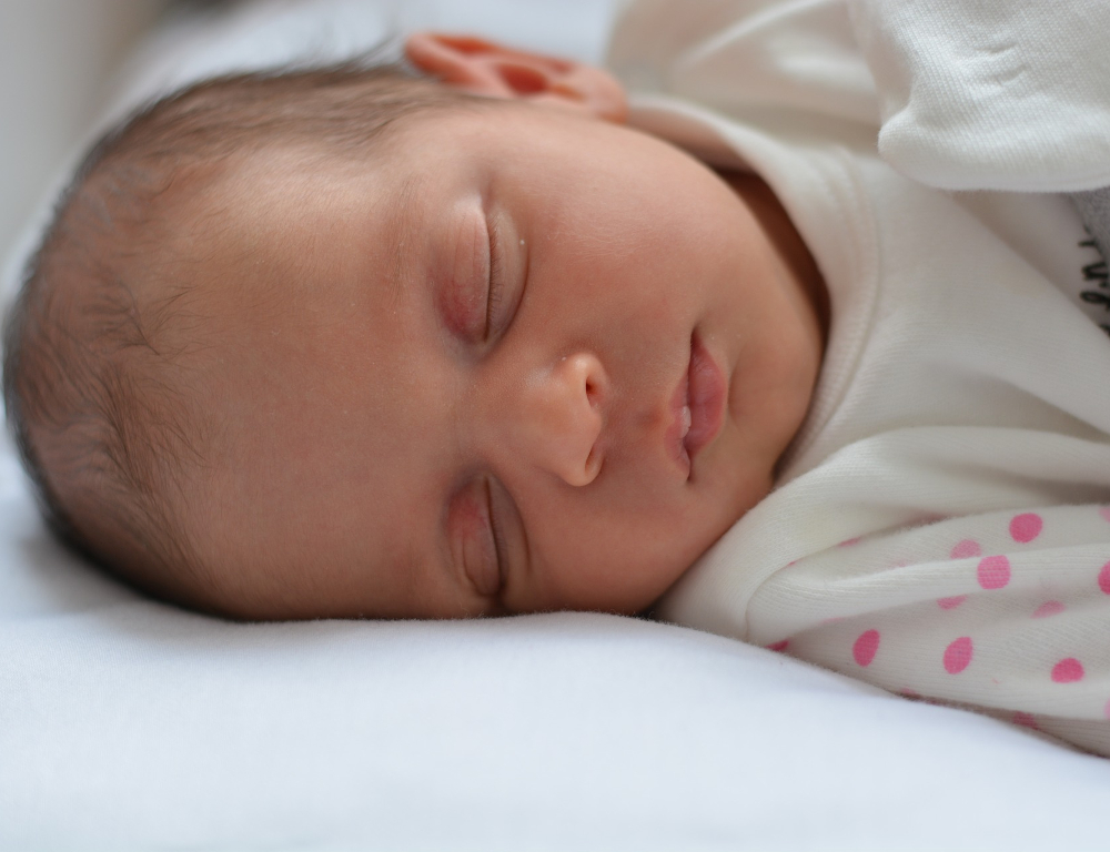 Close-up of a baby laying down sleeping, on a white sheet. The baby's white outfit has pink polka dots on the arms.