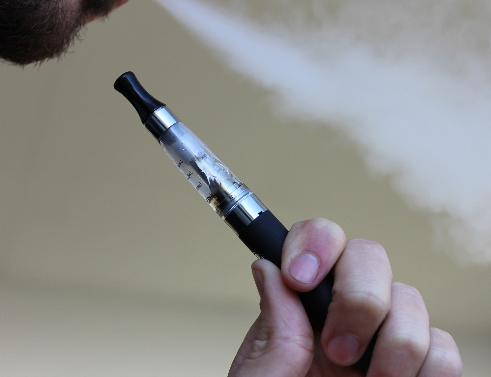 Close-up of an e-cigarette being grasped by a hand. At the top of the photo, a mouth is exhaling vapor.