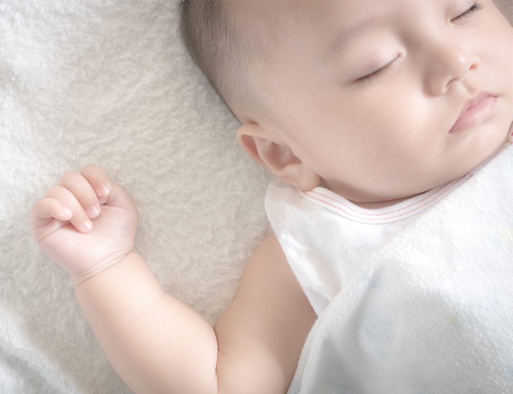 Close-up of an infant sleeping on its back on a white blanket, wearing a light pink shirt.