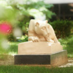 The Penn State Nittany Lion statue is seen in Penn State College of Medicine's outdoor courtyard. The statue is seen at the right of the photo in focus, framed by trees and pink flowers to the left and above and grass below.