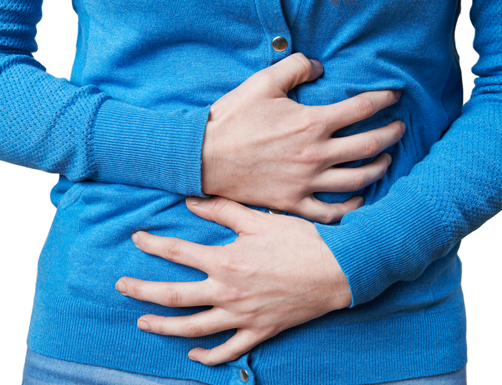 A close-up of the mid-section of a woman wearing a blue long-sleeved sweater, as she grasps her stomach and intestinal regions with both hands.