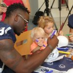 A Penn State football player sits at a table with two small children. The child in the foreground is drawing on a white mini-football helmet with a marker, along with the player.