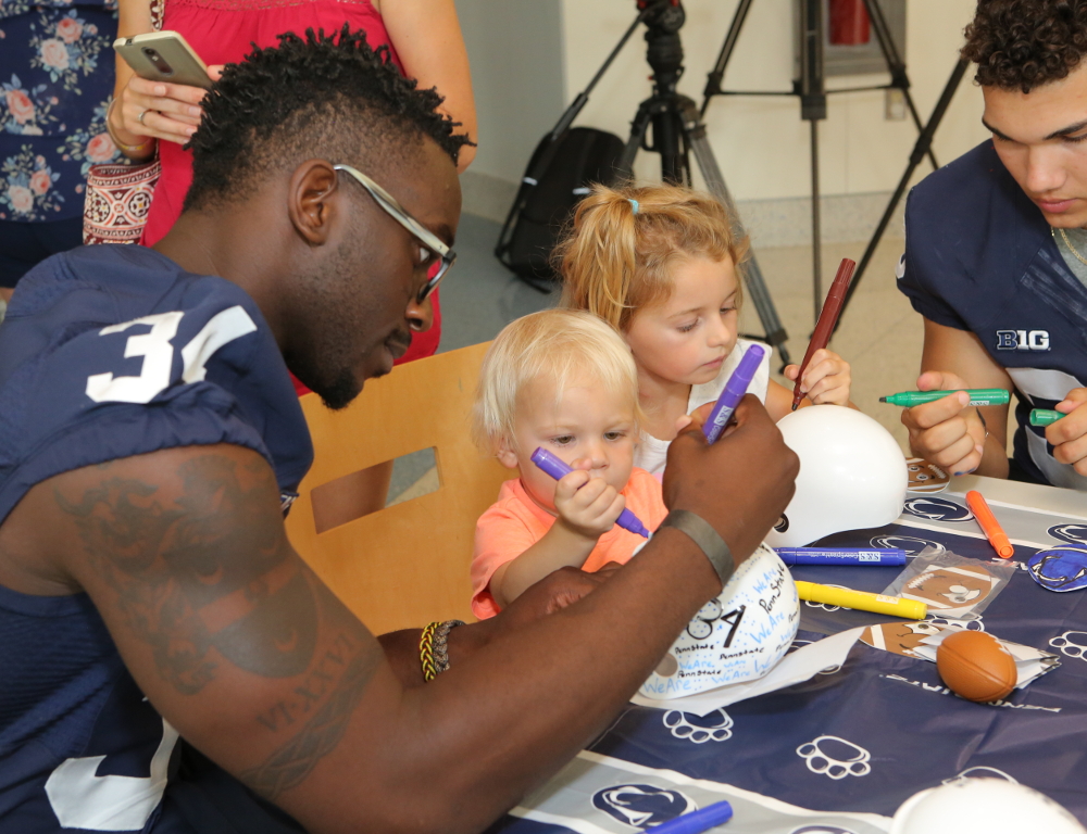 A Penn State football player sits at a table with two small children. The child in the foreground is drawing on a white mini-football helmet with a marker, along with the player.