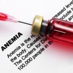 A closeup of a syringe, the needle of which is penetrating the top of a small red bottle. The backdrop is a sheet of paper containing the word "Anemia" and an extensive description of the condition. Only some of the words are readable.