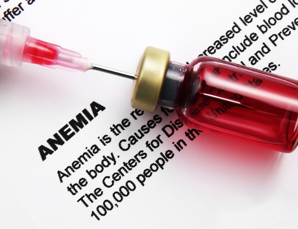 A closeup of a syringe, the needle of which is penetrating the top of a small red bottle. The backdrop is a sheet of paper containing the word 
