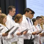 Approximately eight people wearing white doctor coats stand in a row and look down at a paper off of which they are reading in unison.