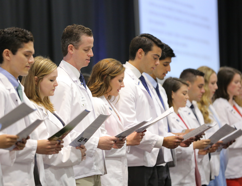 Approximately eight people wearing white doctor coats stand in a row and look down at a paper off of which they are reading in unison.