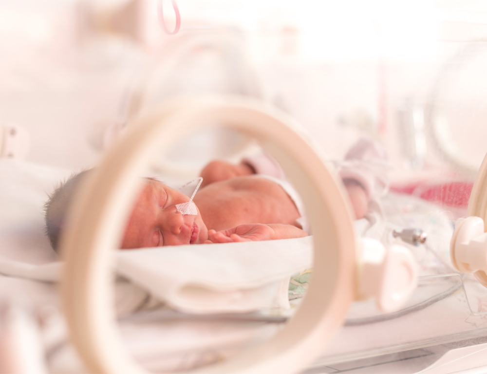 A baby lays on its back in a NICU isolette, its head turned toward the camera, which peers through a hole in the side of the unit.