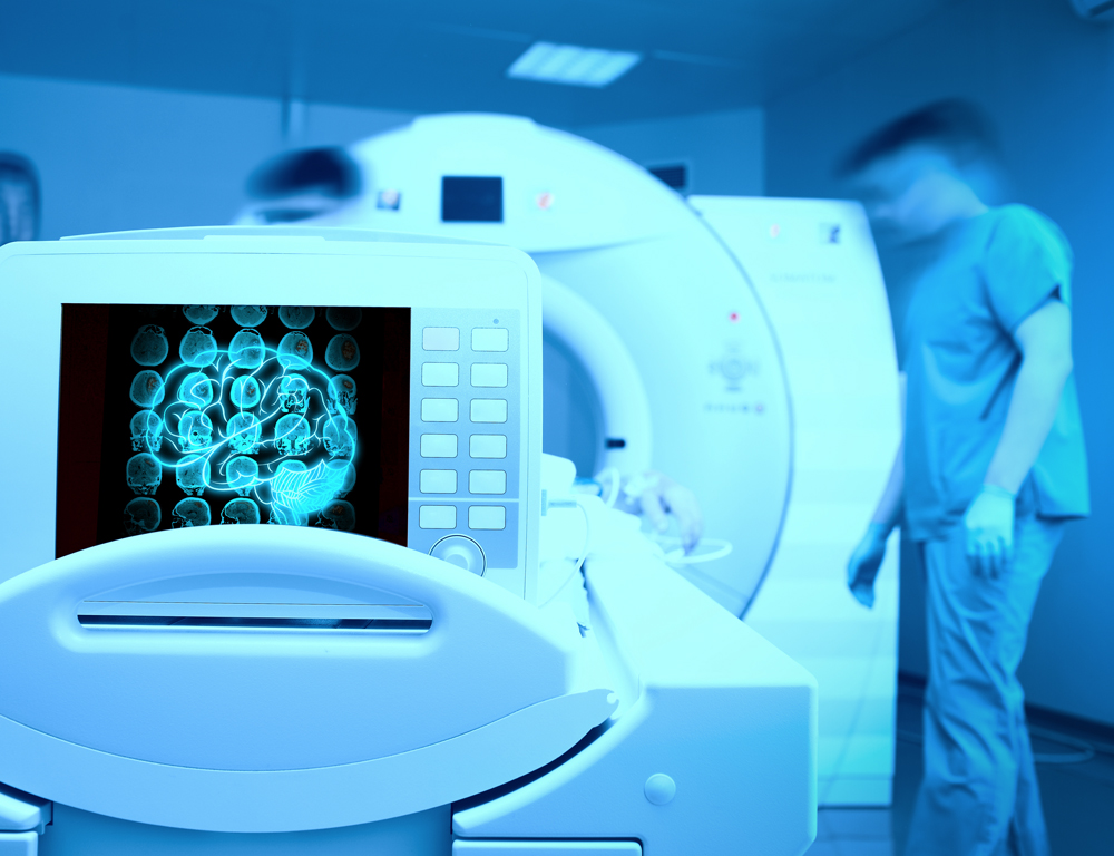 A medical monitor bearing the image of a brain is in the foreground, sitting on a larger piece of medical scanning equipment. The blurred likenesses of two individuals can be seen in the background.