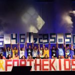 Several people stand on a stage and hold up numbers comprising the figure $6,461,295.50. In front of them are large red letters reading "FOR THE KIDS."