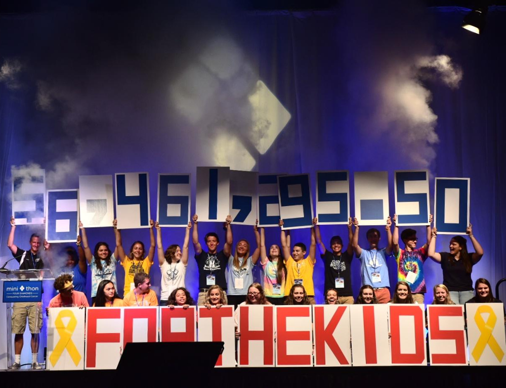 Several people stand on a stage and hold up numbers comprising the figure $6,461,295.50. In front of them are large red letters reading 
