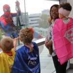 Three young children wearing capes, including a girl being held by her mom, look out a window at Spiderman, who is rappelling down the outside of the building.