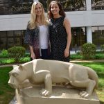 Caitlin McMenamin, left, and Haley Nation, right, recently completed their final thesis dissertations and graduated from Penn State College of Medicine's Anatomy PhD program. The two are pictured in an outdoor courtyard at the College of Medicine, with a Penn State Nittany Lion statue in front of them, a building and a grassy area with shrubs in the background.