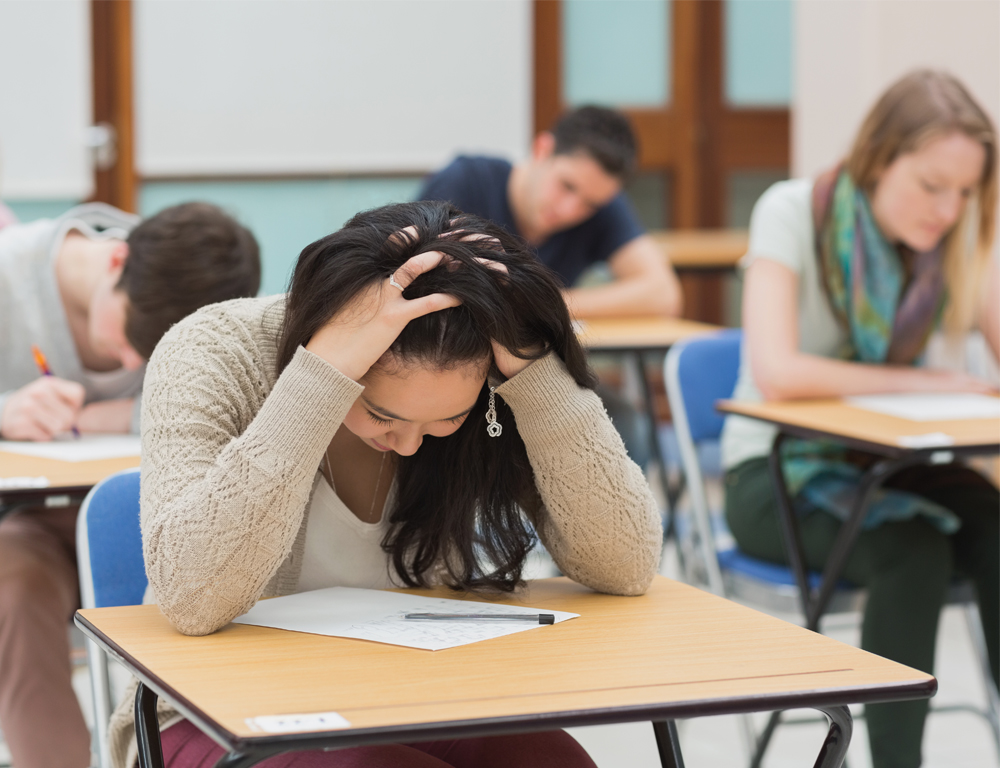 A teenage girl sits at a school desk, holding the top of her head with her hands, looking down at a paper on the desk. Three other students are in the background, slightly out of focus.