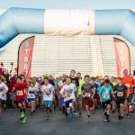 A group of children and adults in running clothes run through a large blue inflatable arch, underneath which there are signs reading 'start' and 'finish.'