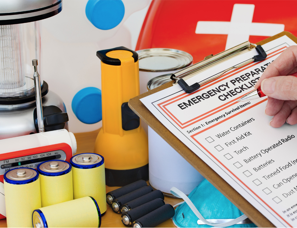 A hand holds a pen above a clipboard bearing a sheet reading Emergency Preparation Checklist. Items such as batteries, a flashlight and a radio are in the background.