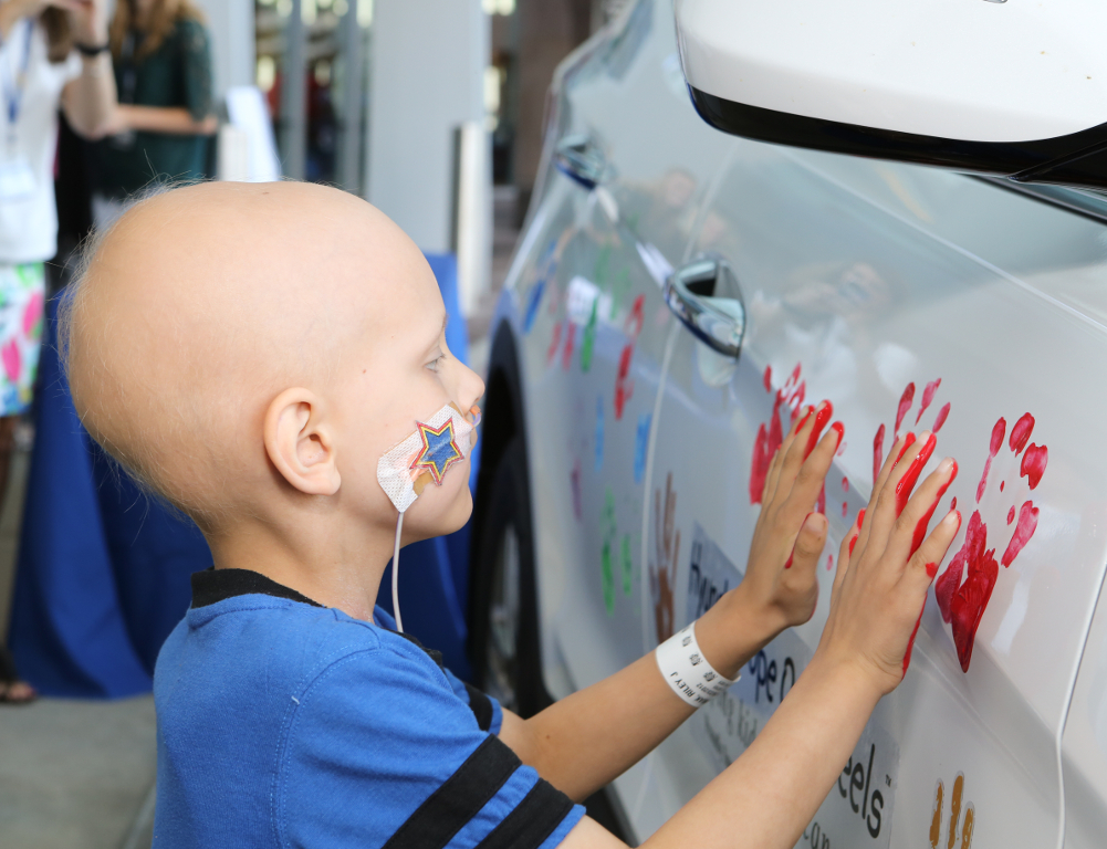 A young child in a blue shirt, and with a hospital bracelet and nasal feeding tube, places both hands “ which are coated in red paint “ on the side of a white car.