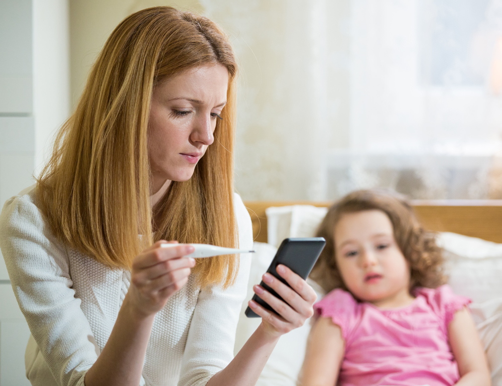 In the foreground, a woman holds a digital thermometer and looks down at her phone, in the other hand. In the background, a young girl in a pink shirt lies in bed.