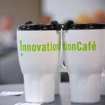 White travel mugs displaying the bright green Innovation Cafe type logo are seen sitting on a table in focus at the front right of the photo. Out of focus behind them, a crowd of people is seen attending an Innovation Cafe event.