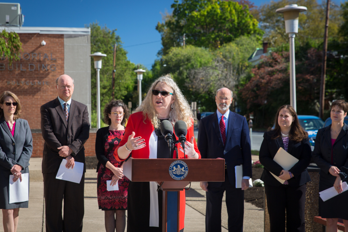 A woman speaks at a lectern as several people stand in a line behind her, looking on. A brick building and some trees are in the background.