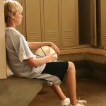 A young boy sits on a concrete bench in a locker room, surrounded by lockers. He holds a basketball in his lap and leans back against a locker, his eyes closed.
