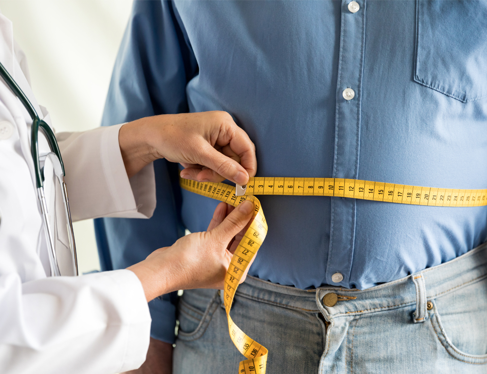 A physician in a white coat and a man in a blue button-down shirt and blue pants are seen from the torso down. The physician is holding a tape measure around the man™s waist.