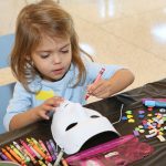 A young girl sits at a table, drawing on a white plastic mask with a red crayon. Other crayons and craft supplies are on the table near her.