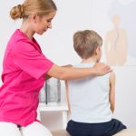 In a clinical exam room, a woman sits beside a young boy on an exam table. Her hands are on his shoulders and she appears to be looking at the boy™s spine.