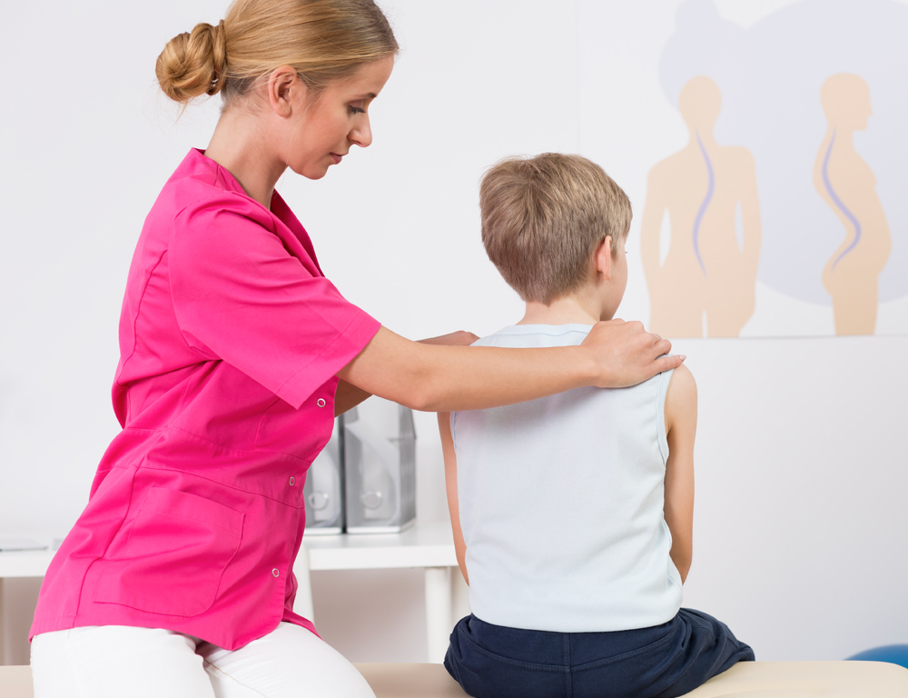 In a clinical exam room, a woman sits beside a young boy on an exam table. Her hands are on his shoulders and she appears to be looking at the boy™s spine.