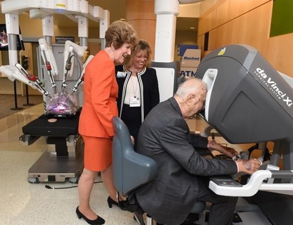 A man sites at the controls of a da Vinci Xi surgical system. Two women stand behind him, looking on.