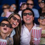 Two parents and two children sit together in a movie theater. All four wear black-rimmed 3-D glasses. They are looking at the screen and smiling widely. Other people are in the background, out of focus.