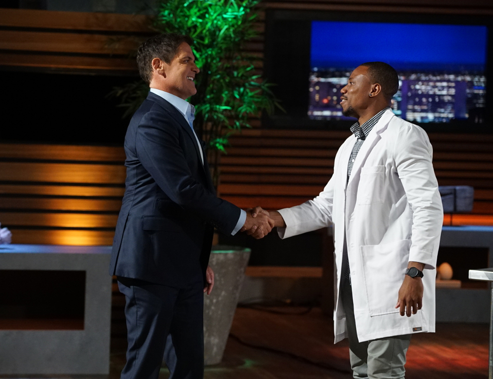 A man in a dark suit (at left) and a man in a white lab coat shake hands and smile. In the background is a TV set with wood paneling and a tall green plant.