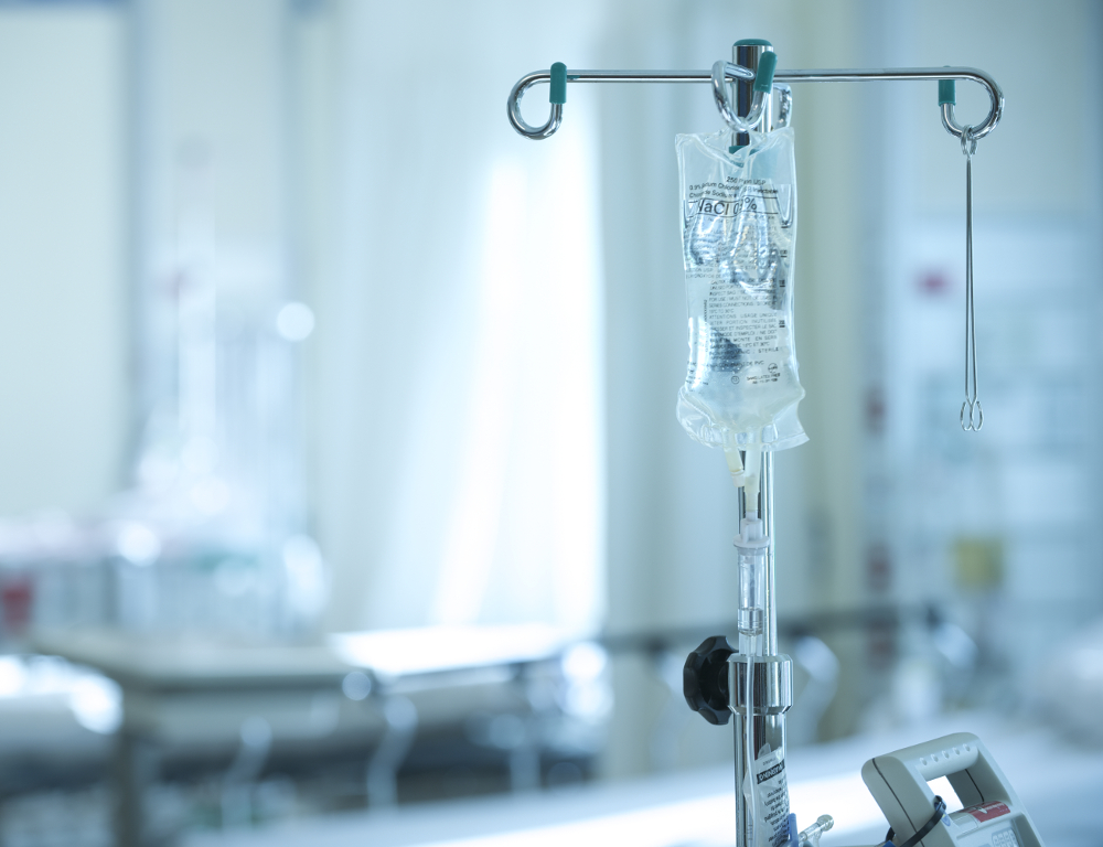 An IV bag hangs off of a metal IV pole. Various components of a hospital room are in the background, slightly out of focus.