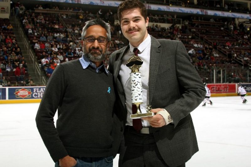 Two men pose for a photo, looking at the camera. The man on the right holds a small trophy. They are standing on an ice rink, where some young hockey players and fans are seen in the background.