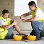 A man in a yellow construction vest sits on the ground against a wall, holding his knee in apparent pain. Another main in a construction vest kneels next to him, preparing to apply a bandage.