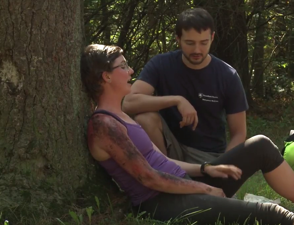 A woman in a purple tank top and obvious (mock) injuries to her right arm sits and leans against a tree. A man in a blue t-shirt kneels beside her.