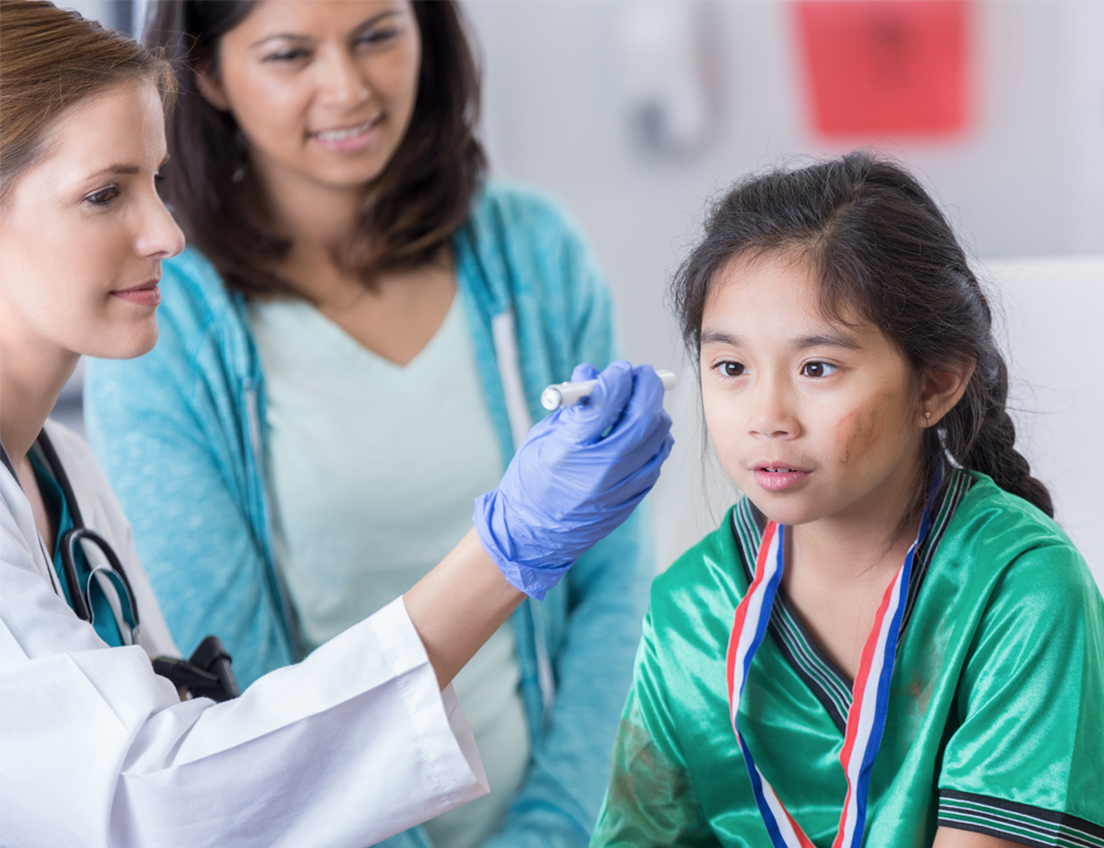 A young child in a green soccer jersey looks straight ahead into the beam of a small flashlight being held by a physician's gloved hand. The girl's mom looks on in the background.