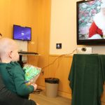 A young girl sits in her mom™s lap. The girl looks up at a TV screen bearing an image of Santa Claus.