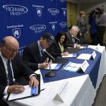 Four people - three men and a woman - wearing business attire sit at a table, each signing a document. A background with the Penn State Health and Highmark Health logos is behind them.